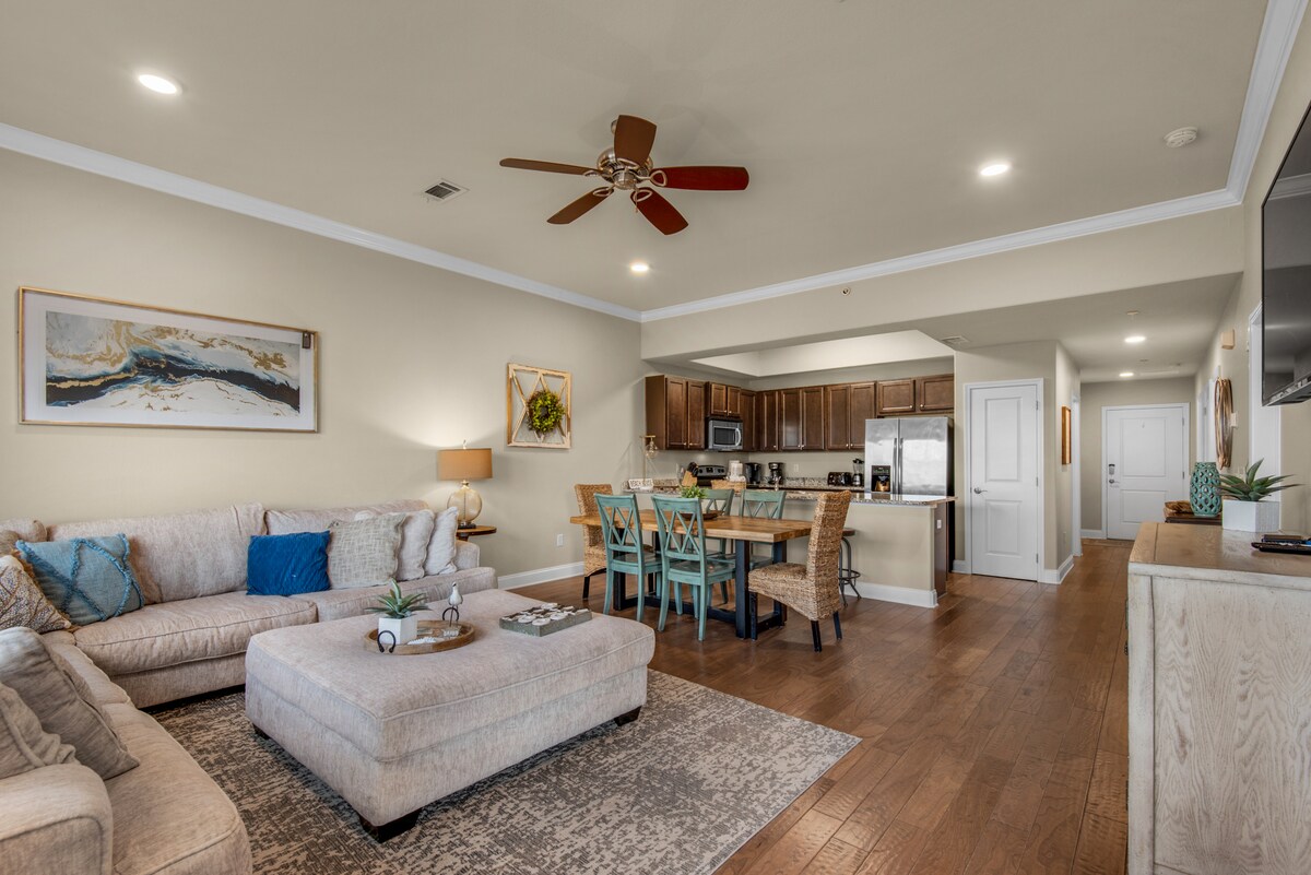 The living area features a large sectional sofa and a comfortable lounge space, complemented by a ceiling fan. A dining area with seating for six is visible, as well as a fully equipped kitchen with dark cabinets and stainless-steel appliances. Natural light fills the room through ample windows.