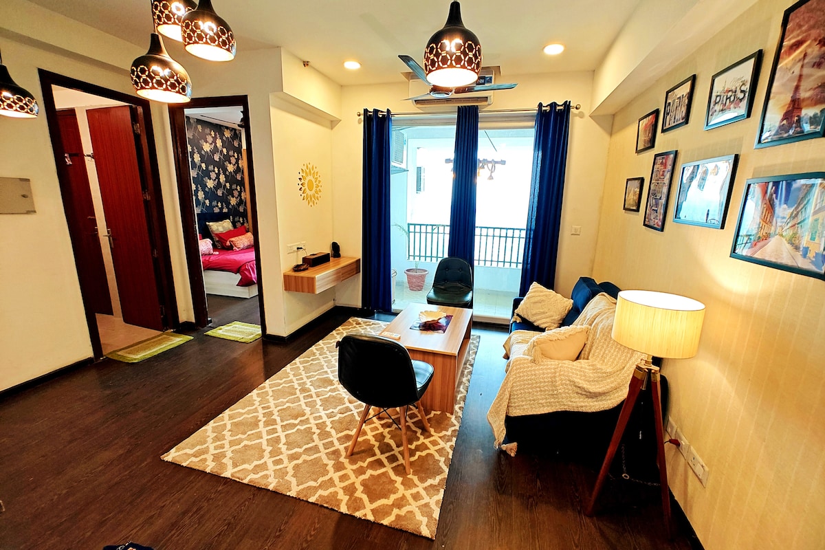 A living area is presented with a comfortable seating arrangement and a wooden coffee table. A patterned rug anchors the space, while large windows are framed by blue curtains, allowing natural light to fill the room. Framed art decorates the walls, providing visual interest.