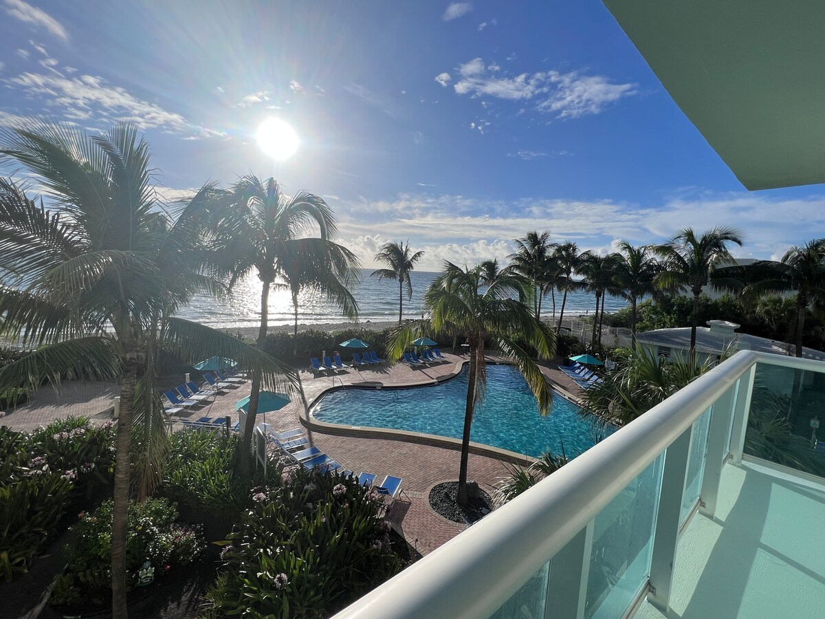 A view from the balcony overlooks a serene beachfront and pool area. Palm trees frame the scene, which is illuminated by the sun reflecting off the ocean. The inviting pool, surrounded by lounge chairs, enhances the resort's tranquil atmosphere.