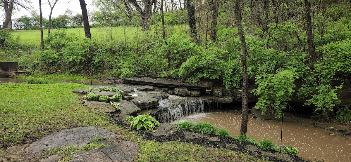 A serene natural setting features a gently flowing spring-fed creek cascading over stones. Lush greenery surrounds the area, with small plants positioned along the water's edge. A wooden footbridge crosses the creek, enhancing the tranquil outdoor atmosphere.