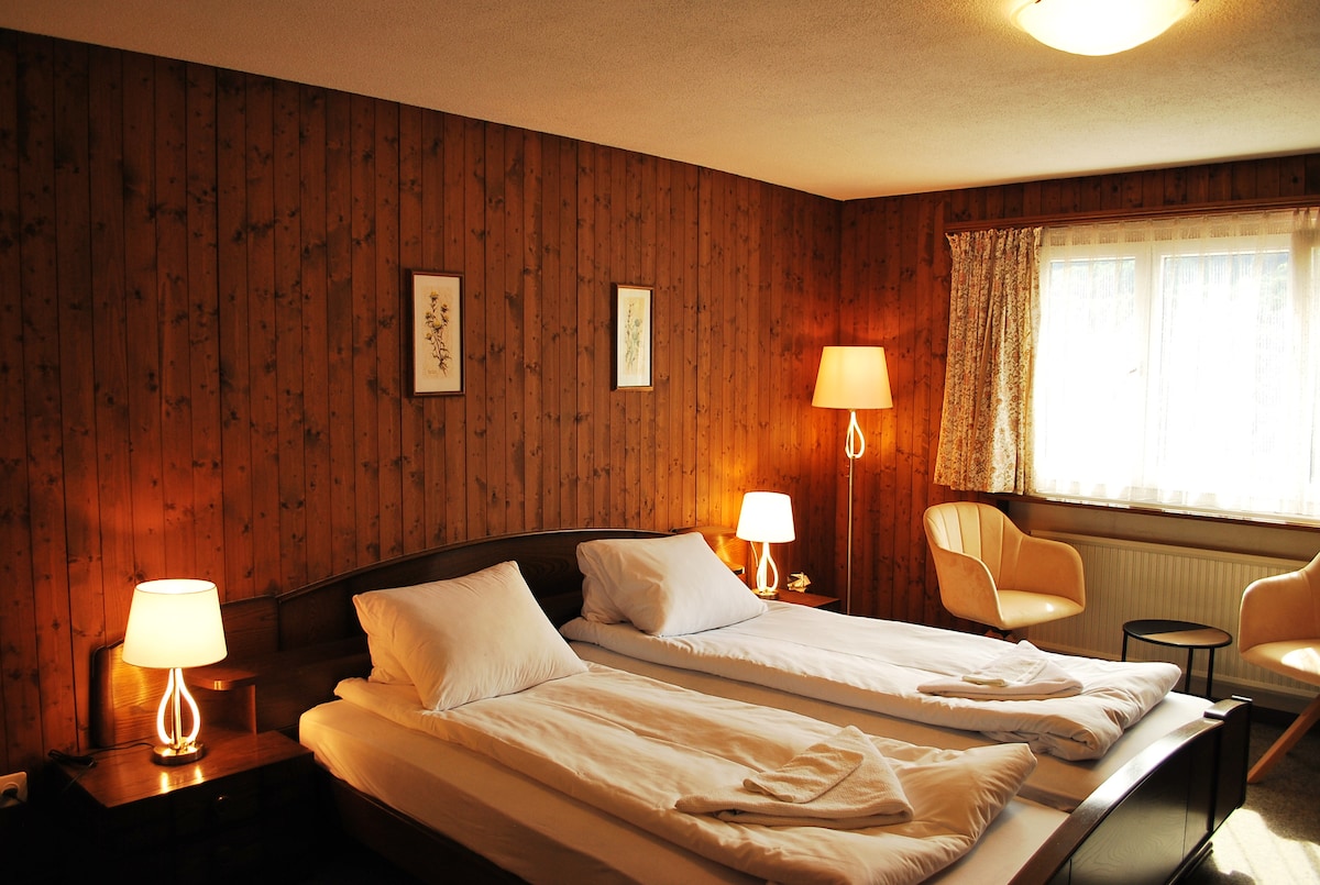 A spacious bedroom is featured with wooden paneling and soft lighting. Two beds with white linens are positioned under a large window, accompanied by two armchairs and a small side table. Framed botanical prints are displayed on the walls.