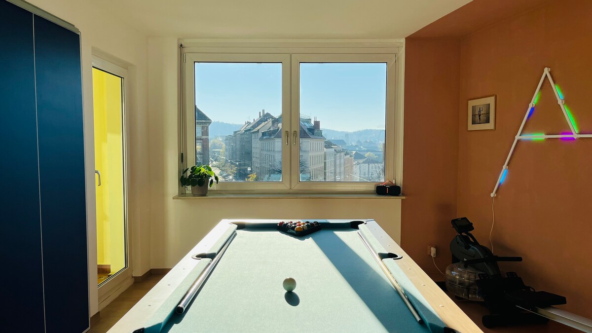 A bright room features a billiard table set against a large window that offers a view of the city. Natural light fills the space, highlighting light-colored walls and a blue cabinet. A plant sits on the window sill, adding a touch of greenery.