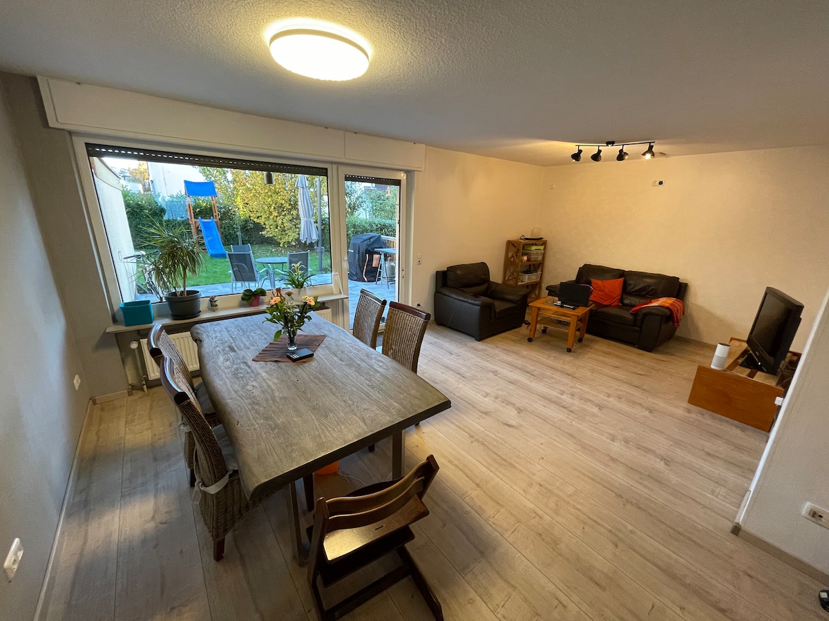 A spacious living area showcases a large dining table surrounded by wooden chairs. A comfortable seating arrangement with dark sofas is visible in the corner. Natural light enters through a large window, offering views of a garden and play equipment outside.
