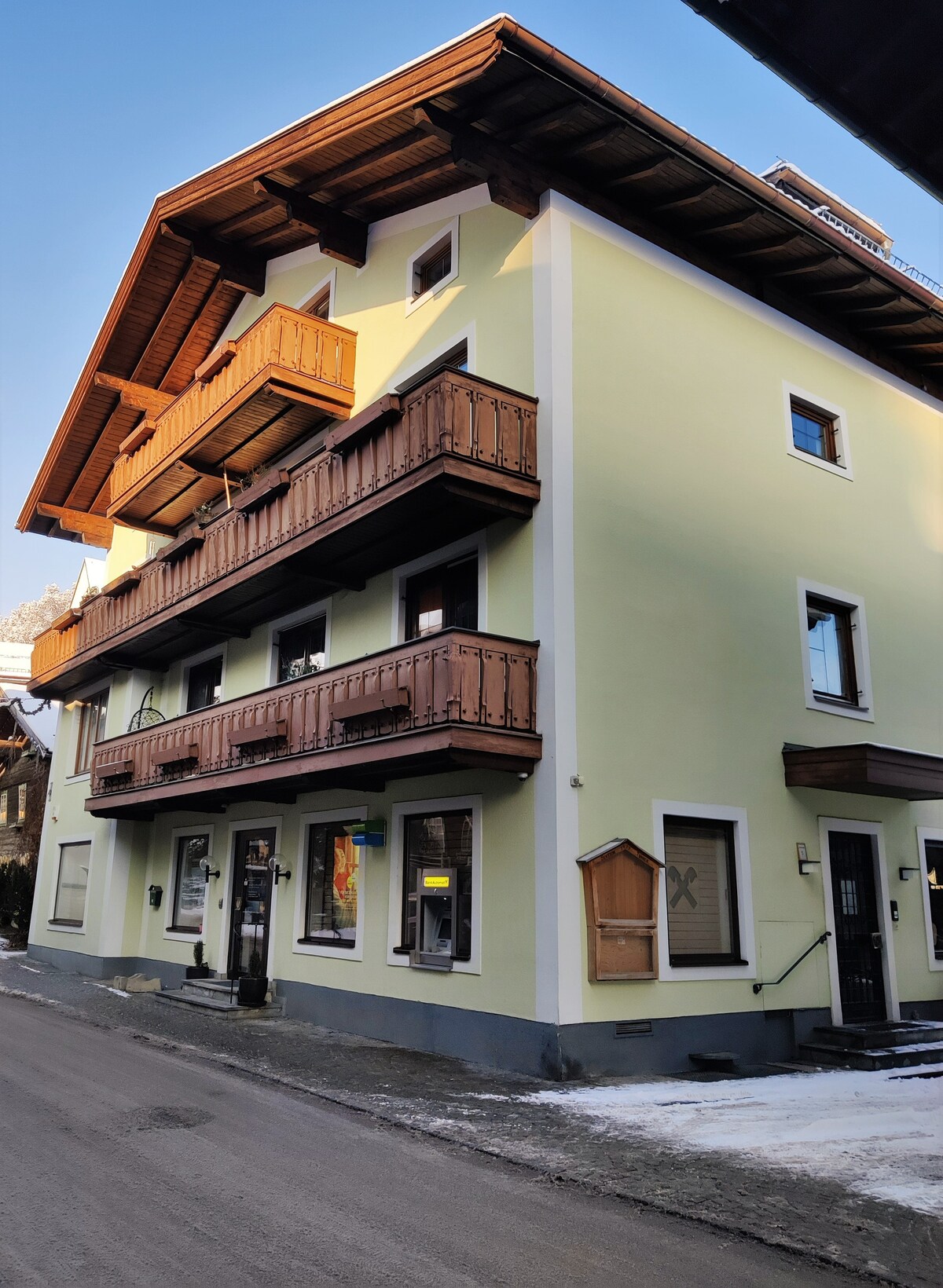 The exterior of a three-story building is depicted, featuring a light green facade and wooden balconies on the upper levels. A well-maintained pathway runs along the building, which includes windows dressed with wooden shutters. The setting suggests a quiet neighborhood ambiance.