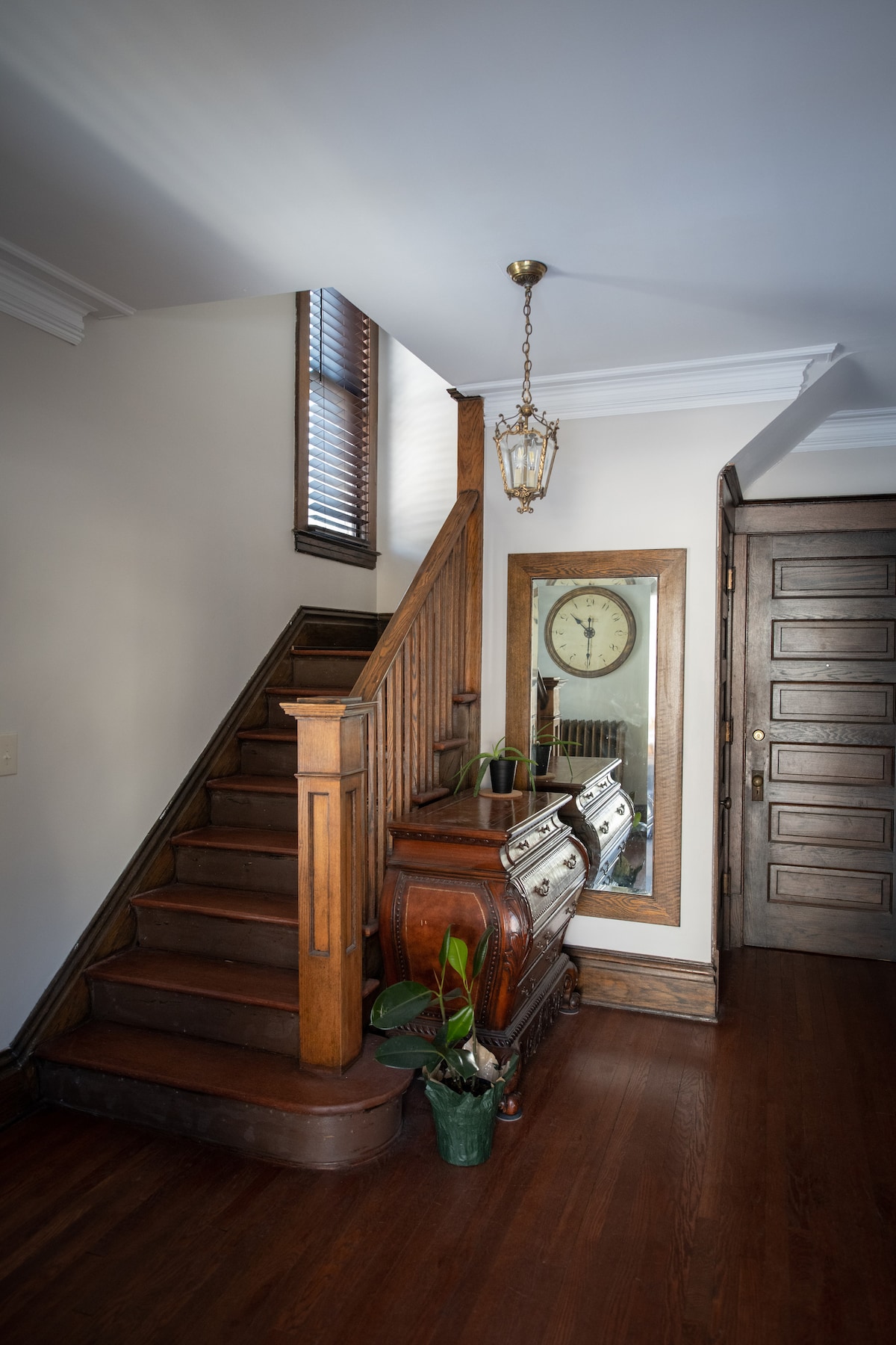 The entrance features a wooden staircase leading to the upper level, illuminated by natural light from a window. An antique mirror reflects the space, while a decorative console table with potted plants adds warmth to the hallway.