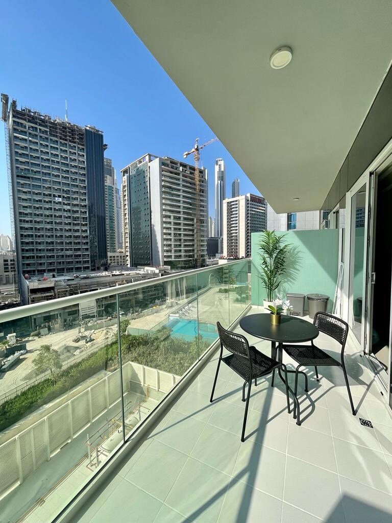 A balcony area is presented, featuring a small black table and two chairs. A partial view of the city skyline is visible, including the Burj Khalifa in the distance. The sunlit space showcases modern glass railing and tiled flooring.