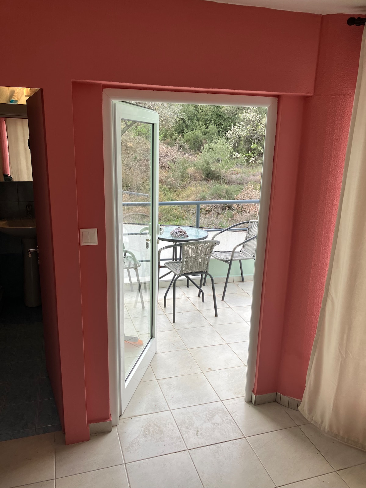 A doorway opens to a balcony area furnished with a table and several chairs. Natural light enters through the glass door, illuminating the pink walls inside. The view beyond shows greenery, enhancing the connection to the outdoor space.
