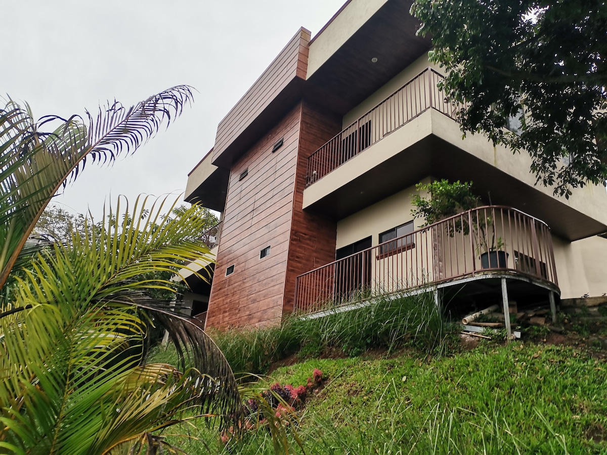 A modern two-story building is situated on a grassy hillside, featuring a blend of wood and concrete elements. The structure includes large windows and a wraparound balcony, surrounded by lush greenery and tropical plants, creating a serene outdoor environment.