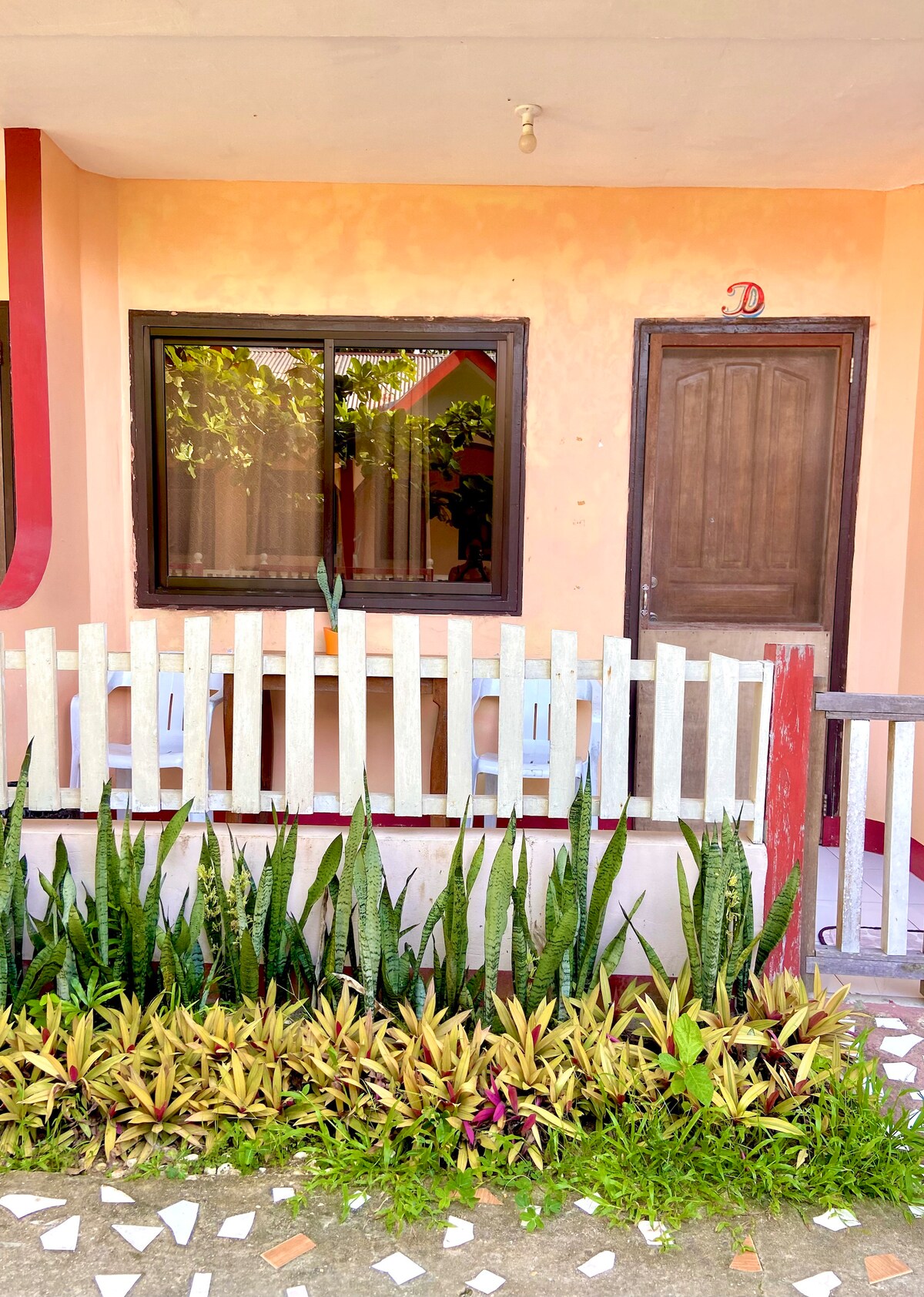 A welcoming entrance features a wooden door framed by a small patio area. Two white chairs are positioned beside a window, allowing natural light to enter. Lush greenery, including tall plants and colorful flowers, borders a white picket fence that adds charm to the setting.