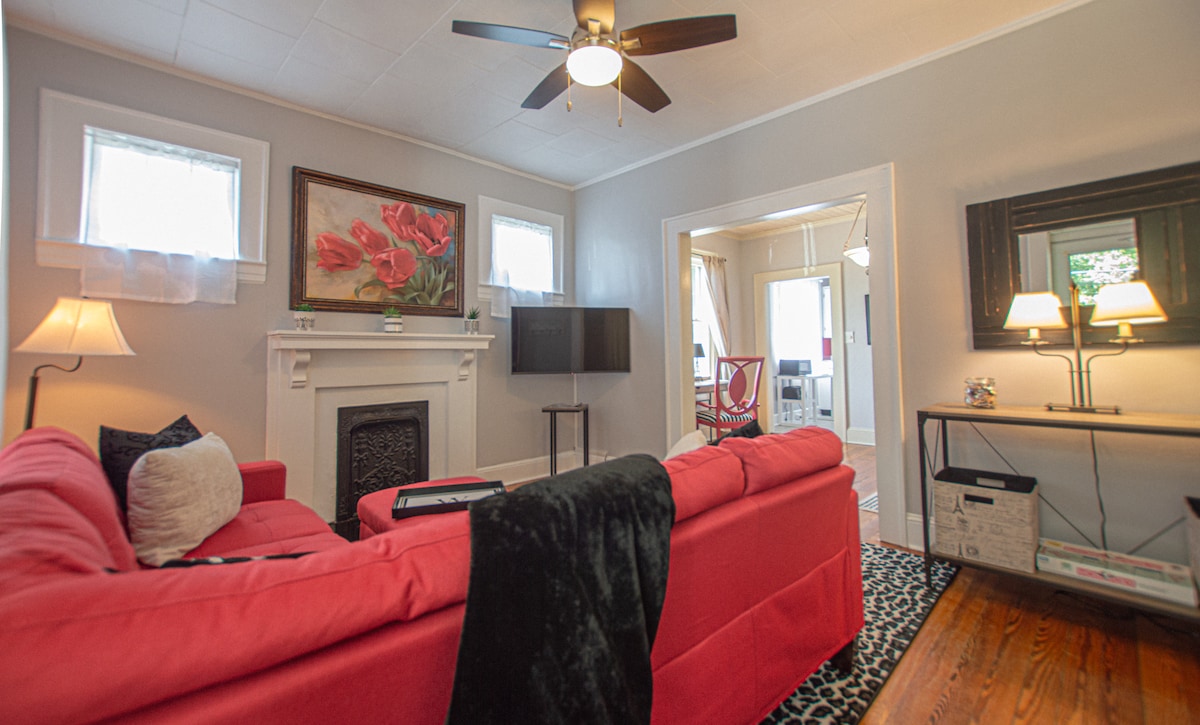 A bright living room is showcased with a red sectional sofa, accented by a black throw. A modern ceiling fan spins gently overhead, while large windows provide ample natural light. A decorative painting of red flowers hangs above the fireplace, complementing the stylish decor.