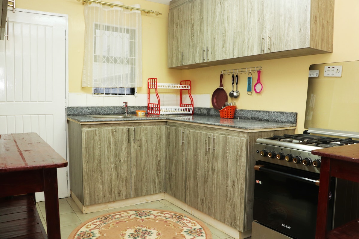 The kitchen area features wooden cabinetry and a granite countertop. A gas stove is positioned beside a stainless steel sink. Kitchen utensils hang on the wall, and a circular rug lies on the tiled floor, complementing the light-colored walls.