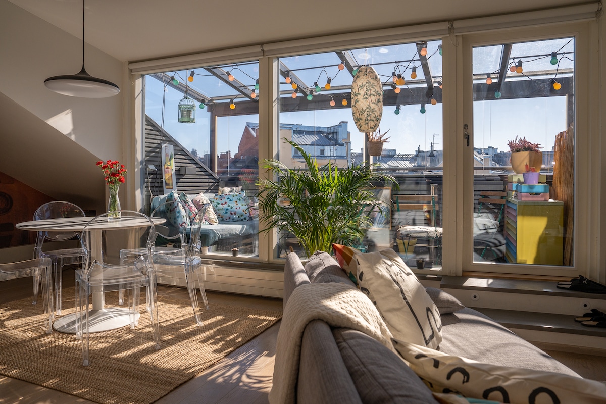 A bright living space features large glass doors leading to a glazed balcony adorned with vibrant decorations and plants. A transparent dining table and clear chairs occupy the area, while a grey sofa adds comfort. String lights create a cheerful ambiance under the roof structure.