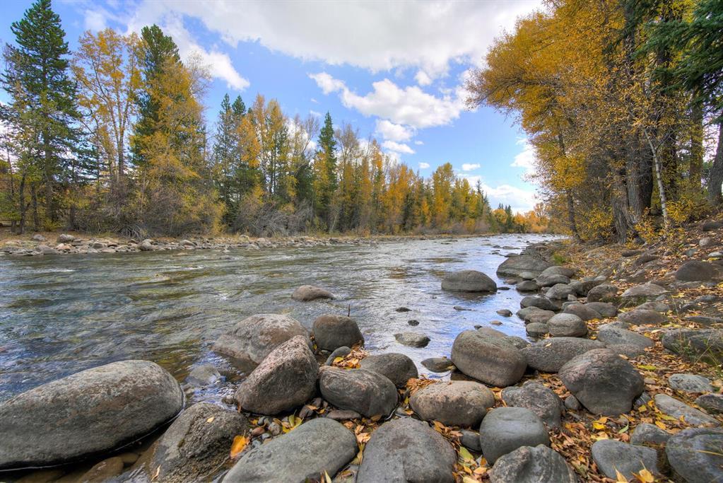 The image captures a serene river scene, with gently flowing water framed by a rich landscape of autumn foliage. Large rocks line the riverbank, surrounded by trees showcasing vibrant yellow and green leaves, under a partly cloudy sky.