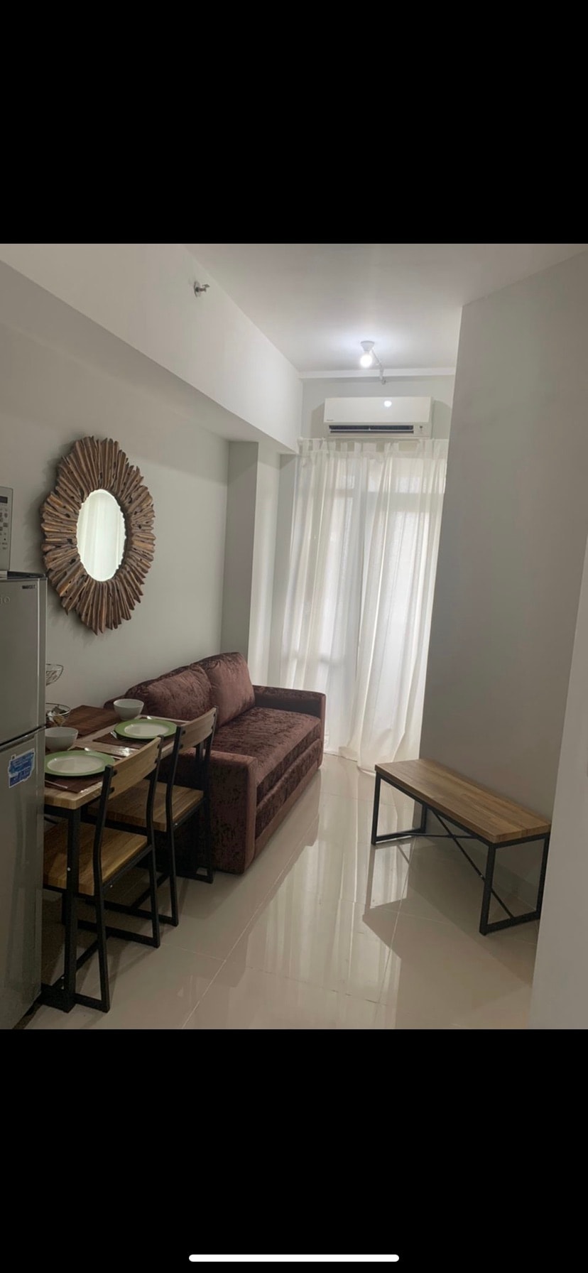 A compact living area is visible, featuring a brown fabric sofa adjacent to a small dining table set for four. A round, decorative mirror adorns the wall, while natural light filters through sheer curtains, brightening the space. A wooden bench provides additional seating.