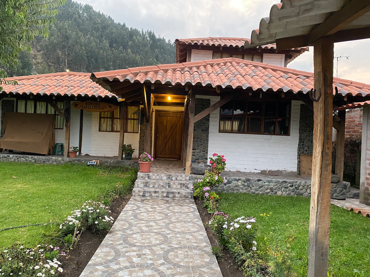 The entrance to a charming accommodation is framed by a landscaped pathway lined with flowers. The structure features a rustic design with a tiled roof and wooden accents, complemented by large windows that allow natural light. Greenery and hills form a serene backdrop.