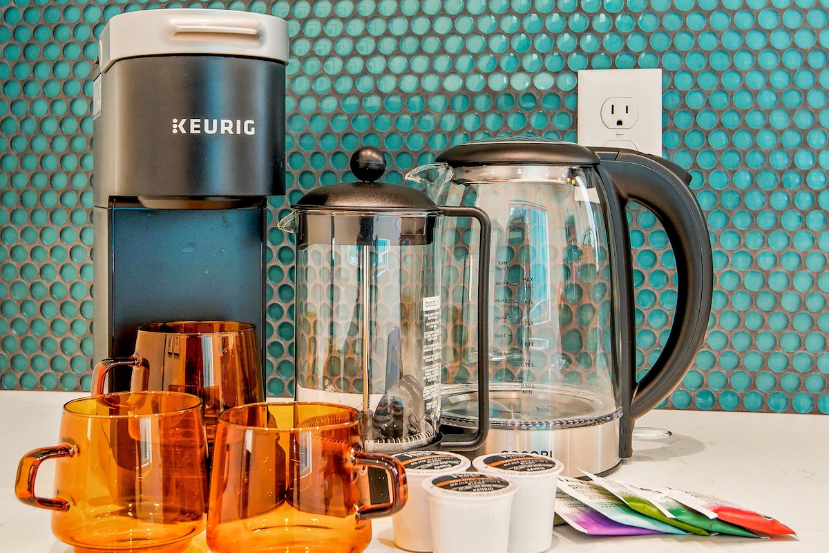 Coffee-making essentials are displayed, including a Keurig machine, a glass French press, and a kettle. Several amber-colored mugs sit in front, accompanied by assorted coffee pods in colorful packaging. The backdrop features a striking blue tiled wall.