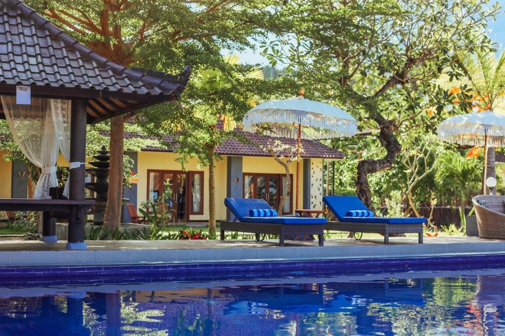 Two sun loungers positioned near a serene swimming pool are surrounded by lush greenery and tropical trees. Traditional Balinese umbrellas provide shade, while colorful flowers enhance the garden's natural beauty. The sun shines on the cozy guest houses in the background, creating a tranquil outdoor setting.
