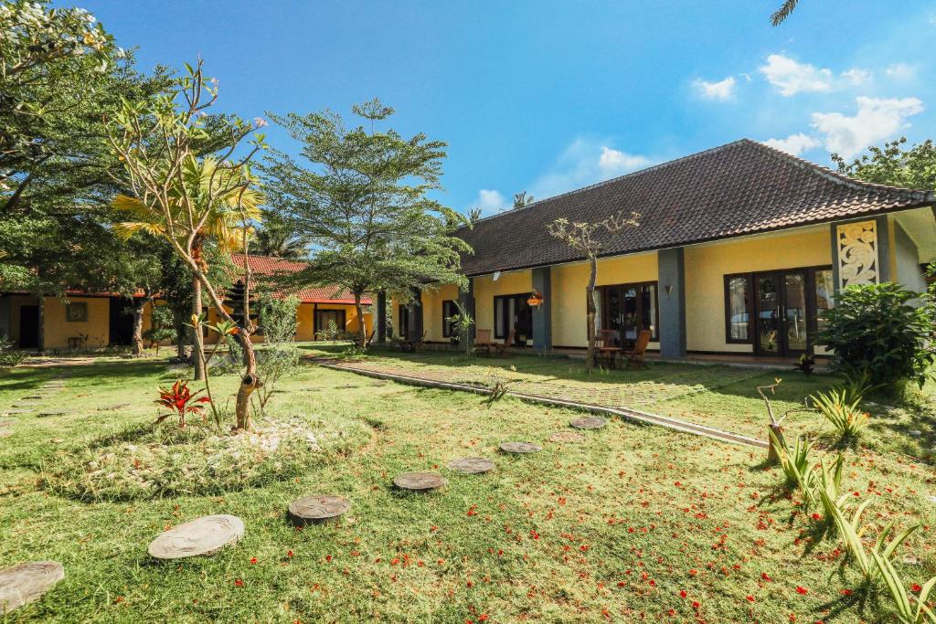 The exterior features a well-maintained garden with green grass and decorative stone pathways. A cluster of tropical plants and trees surround the building, which showcases open-access verandas with large windows. The roofing is a traditional style, complementing the vibrant yellow walls.