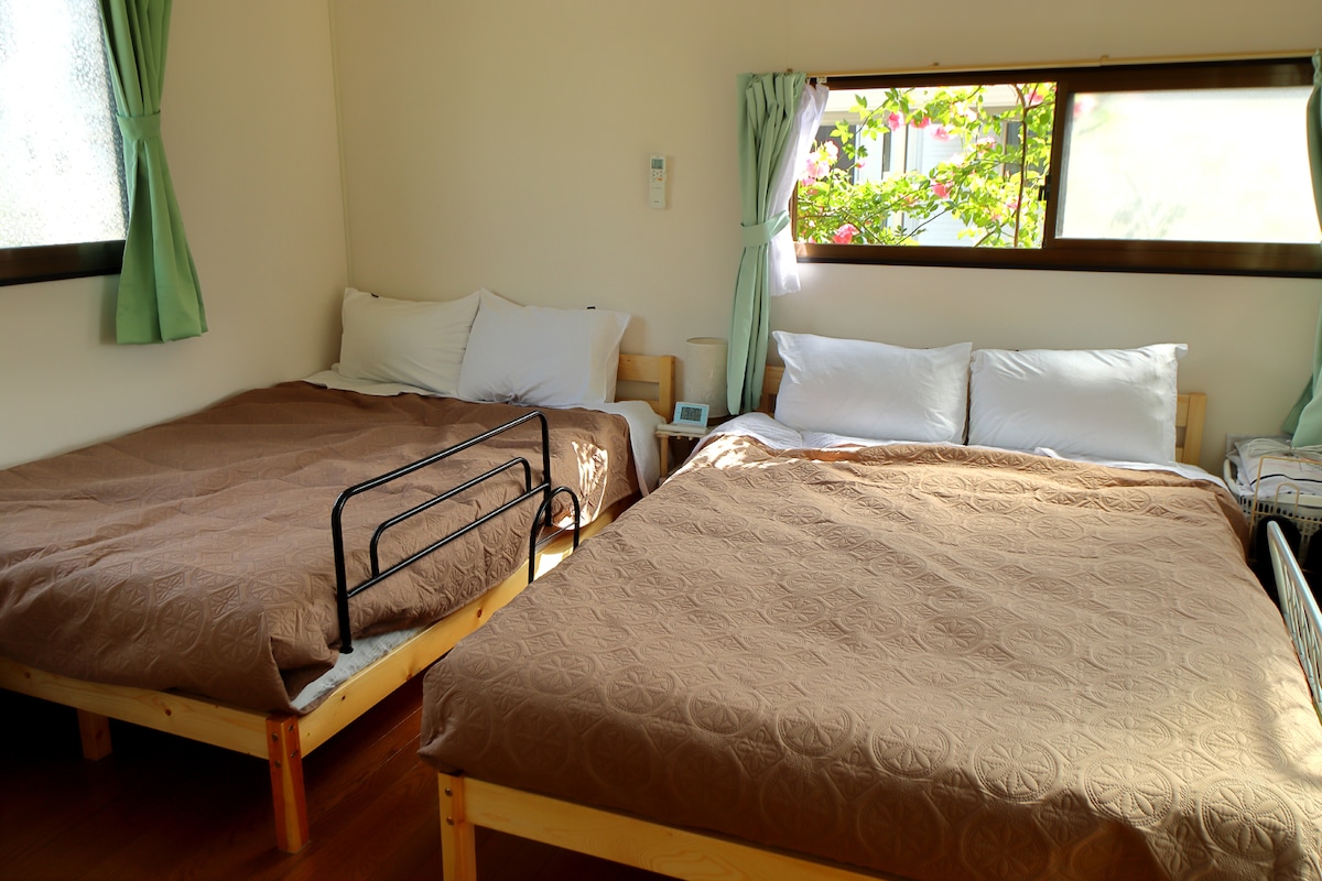 The sleeping area features two spacious double beds adorned with neutral-toned bedspreads. Soft natural light enters through a large window, accompanied by light green curtains. A bedside table sits between the beds, enhancing the room's functionality.