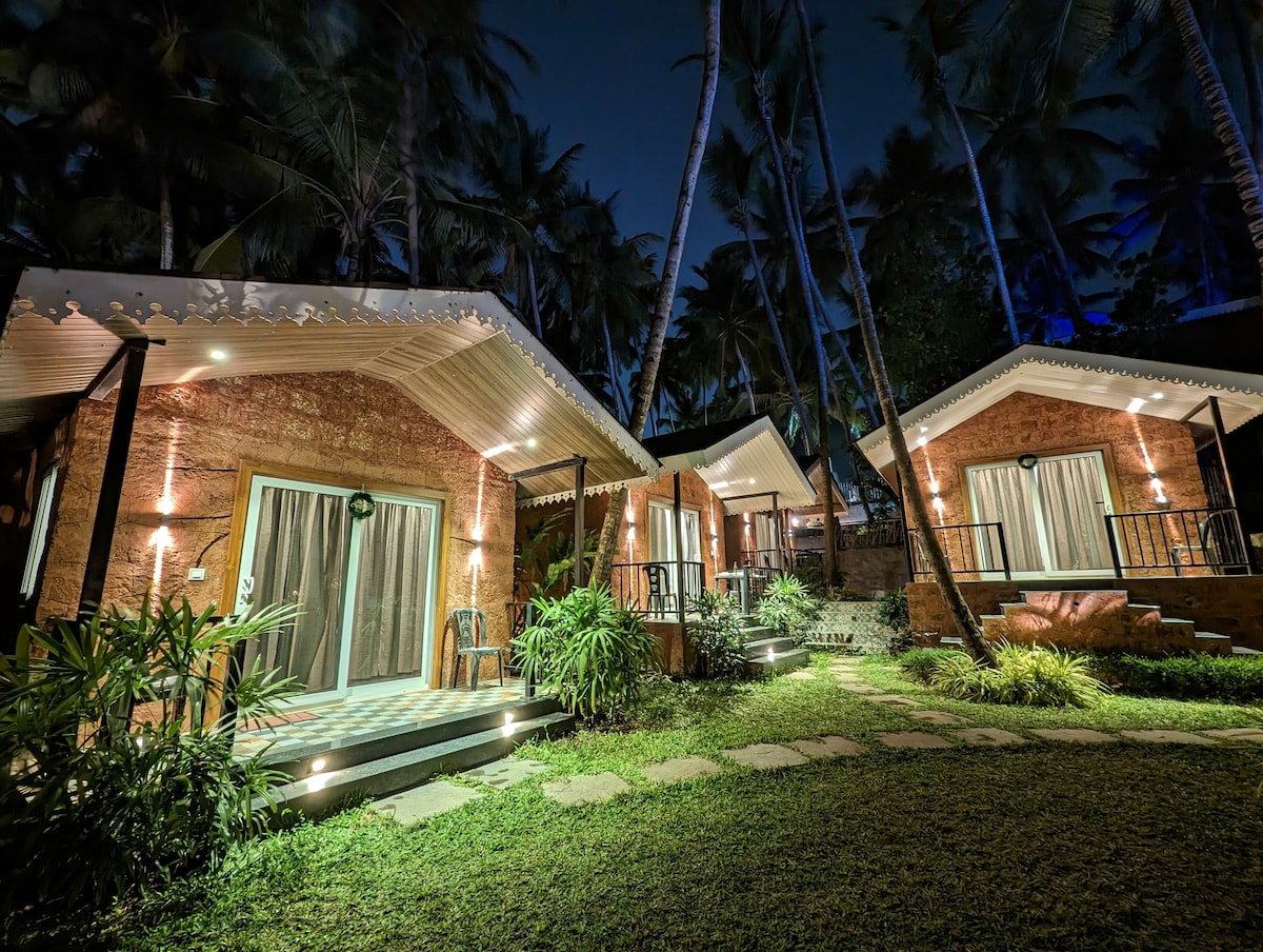 Two charming cottages are set amidst lush greenery, illuminated softly by outdoor lights at night. Each cottage features large glass doors that open to a small porch. The surrounding landscape includes palm trees and well-maintained pathways leading to the cottages.