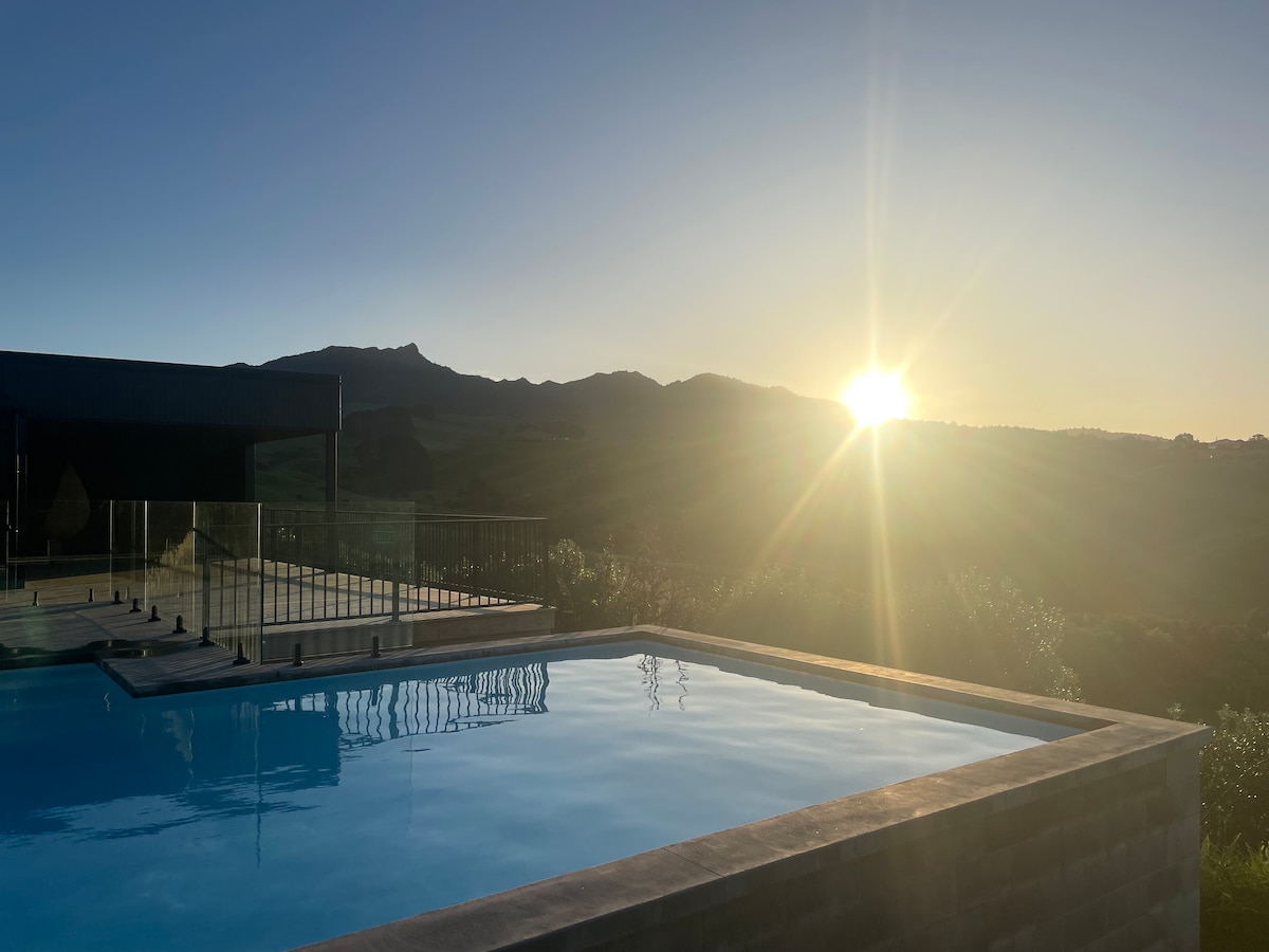 A tranquil pool area is illuminated by the warm glow of the rising sun, reflecting on the calm water. Lush greenery surrounds the pool, with distant mountains visible in the background, creating a serene outdoor setting.