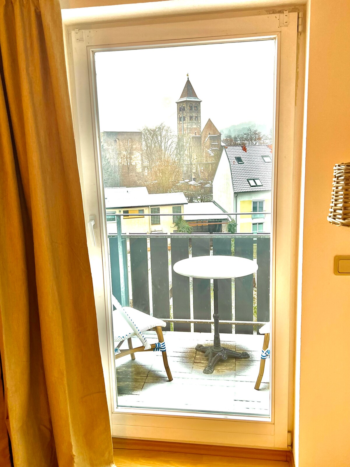 A sliding glass door opens to a small balcony featuring a round table and two chairs. The view includes a historic church tower and surrounding buildings, with soft natural light illuminating the scene.