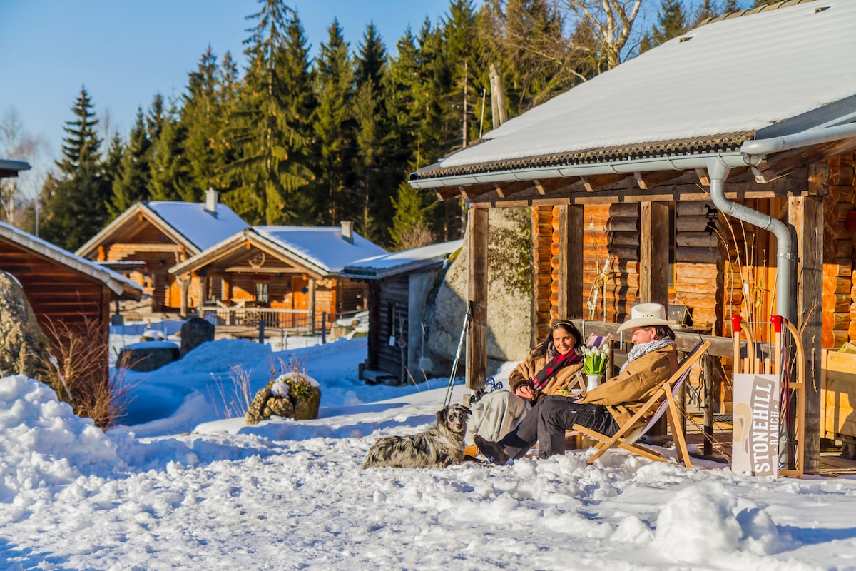 Two individuals are seated in deck chairs on a snow-covered patio. A dog rests nearby. The scene is surrounded by tall trees and several wooden cabins, with clear blue skies above, creating a serene winter atmosphere.
