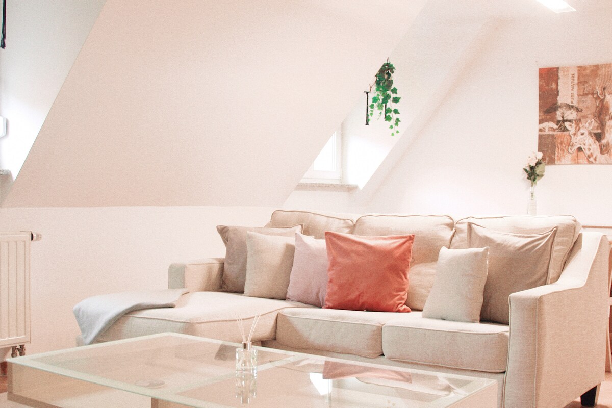 A spacious living area is highlighted by a light-colored sectional sofa adorned with various soft pillows. A minimalist glass coffee table sits in front, and natural light enters through a nearby window. Decorative plants and a piece of wall art enhance the ambiance.