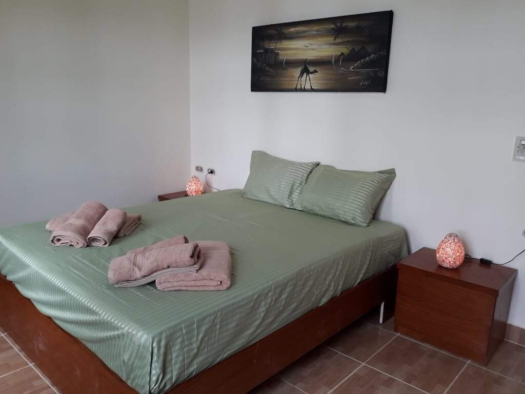 A sleeping area features a spacious bed dressed in soft green linens. Two bedside tables hold decorative lamps that emit a warm glow. Neatly rolled towels are placed at the foot of the bed, and a wall art piece depicting a serene landscape adds visual interest.