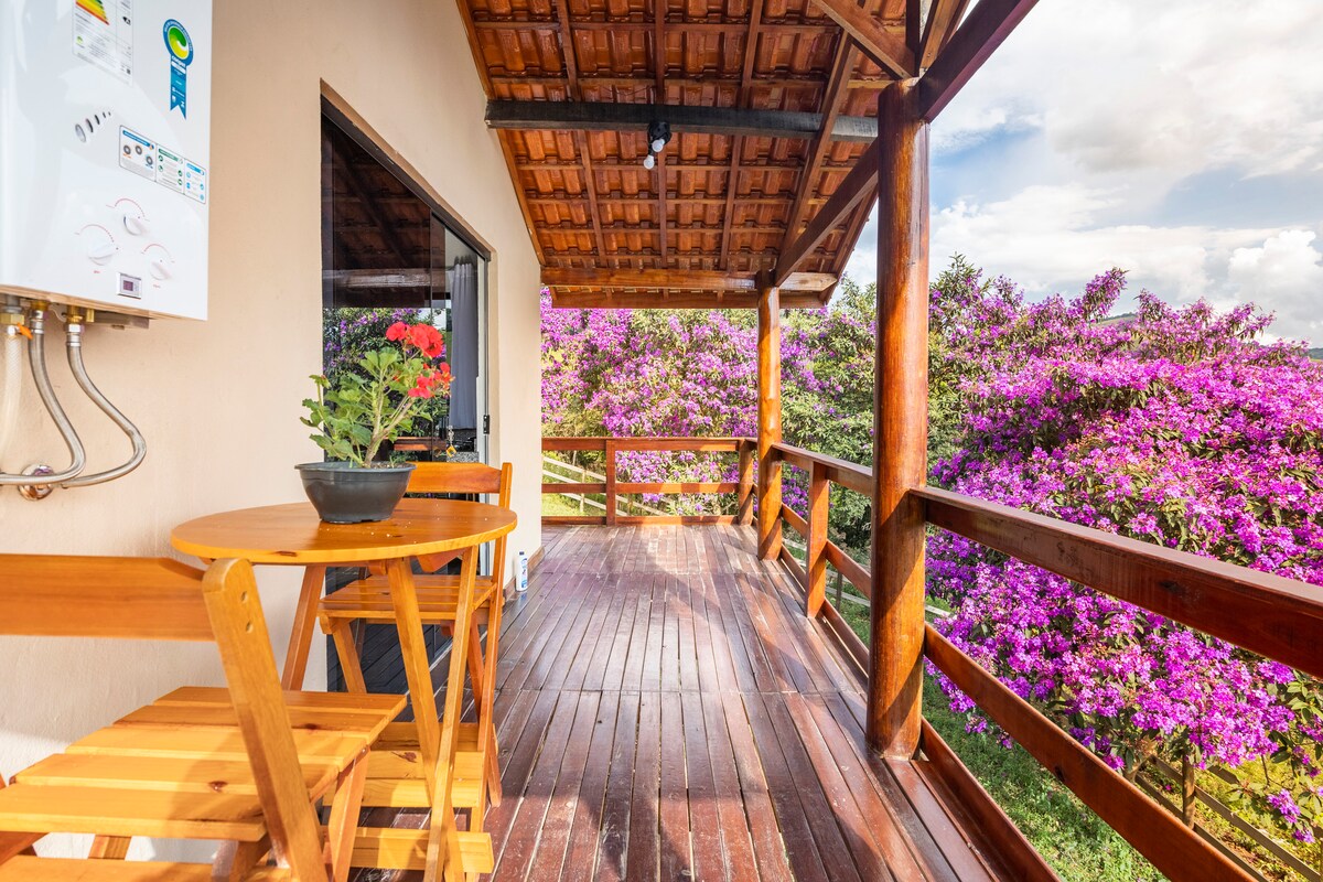 A wooden deck is created with polished flooring and features a small round table and two folding chairs. Vibrant purple flowers are visible in the background, enhancing the natural surroundings. Large glass doors provide access to the interior space.