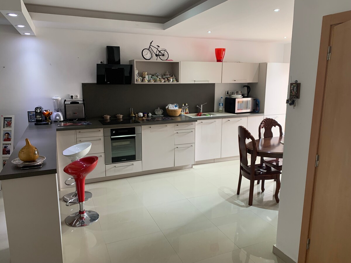 A modern kitchen is showcased, featuring sleek cabinetry and an array of appliances. A dining table with four wooden chairs is present, accompanied by two bar stools at the counter. Bright lighting enhances the clean, minimalist design.