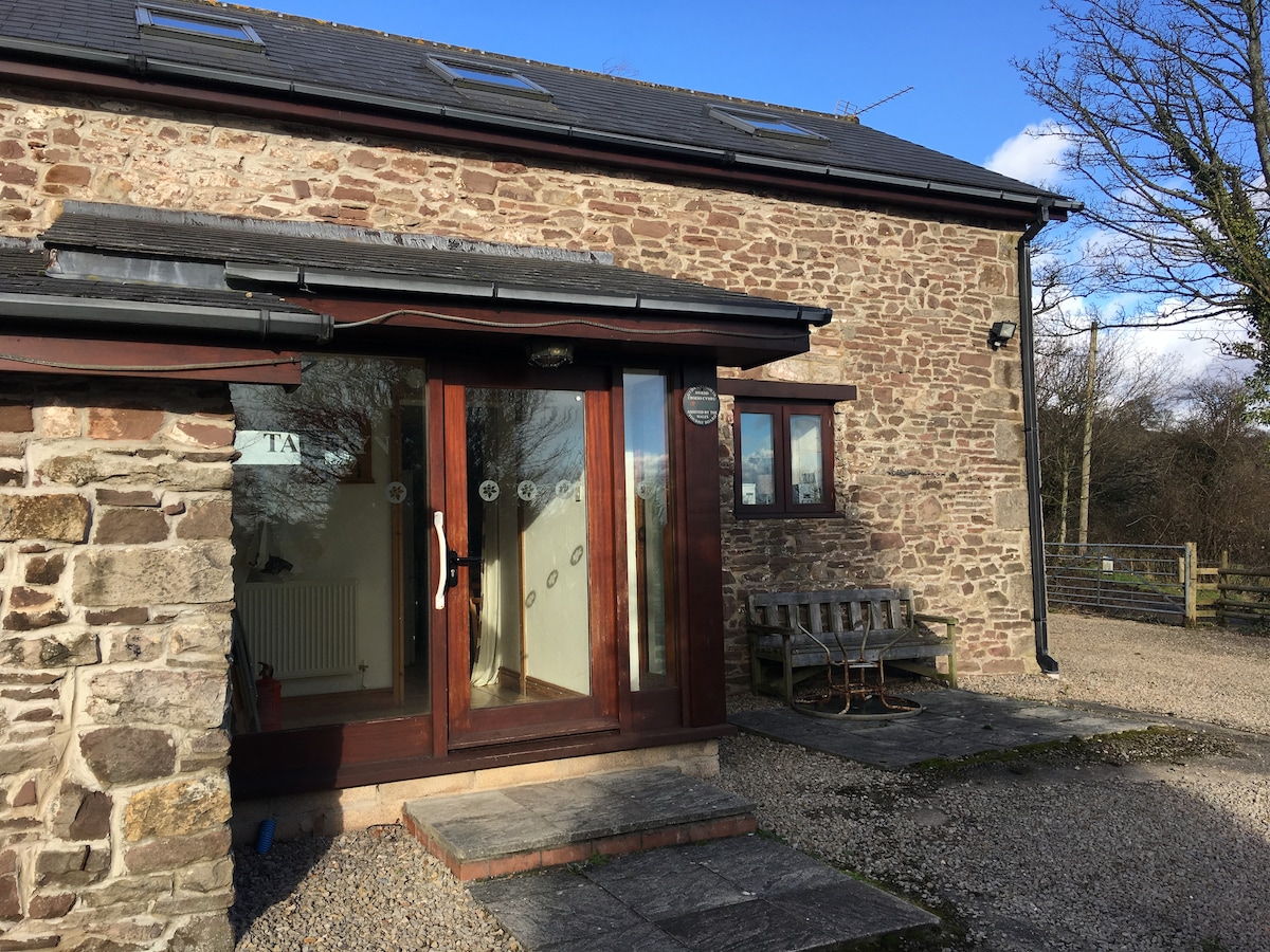 The exterior of a stone cottage is visible, featuring a wooden door with large glass panels. A small stone pathway leads to the entrance, which is flanked by a rustic bench. Natural surroundings include greenery in the background, highlighting a peaceful countryside setting.