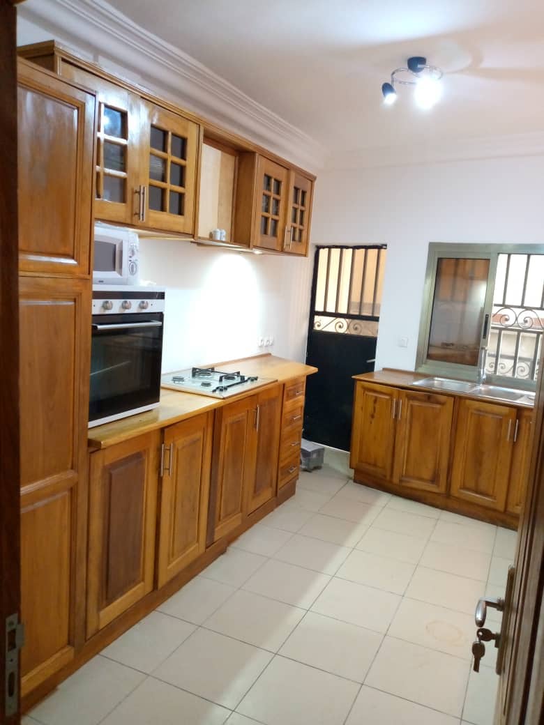 A modern kitchen features wooden cabinetry with glass-paneled doors. The countertop includes a stove, an oven, and a microwave, while a small window allows natural light. The tiled floor is neutral-toned, and the space is equipped with essential appliances for cooking.