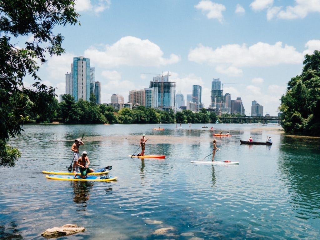 Several individuals are seen enjoying water activities on a lake, with the skyline of downtown Austin visible in the background. Clear blue skies are dotted with clouds, while lush green trees frame the scene, enhancing the vibrant urban landscape.