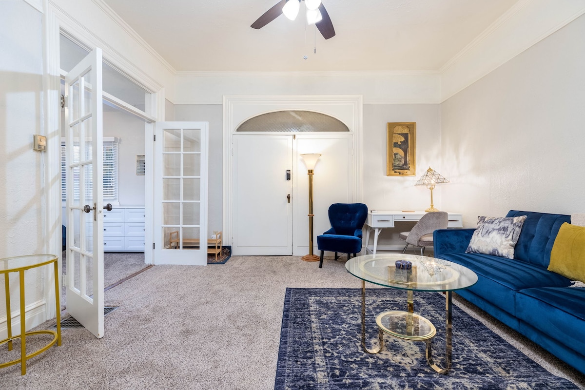 A spacious living area features a plush blue sofa and a glass coffee table atop a decorative rug. Light is provided by a standing lamp, and a cozy armchair adds seating. French doors lead to another room, enhancing the open ambience.