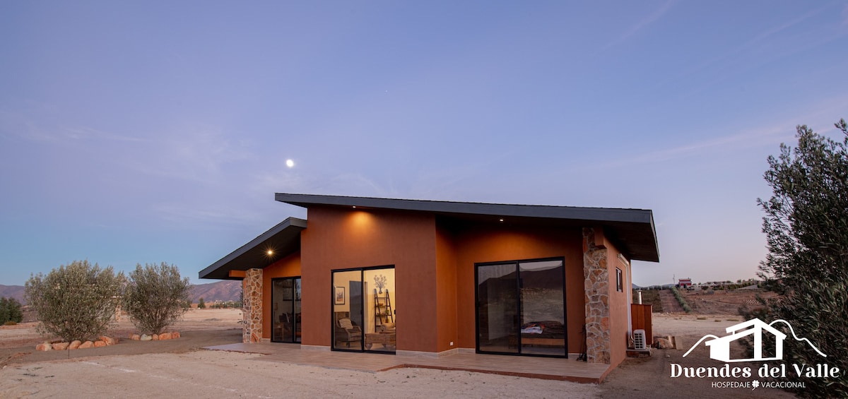 The modern cabin features a sleek design with large glass windows, allowing for abundant natural light. The exterior is complemented by a combination of textured stone and smooth surfaces, set against a serene landscape with distant hills under a clear sky.