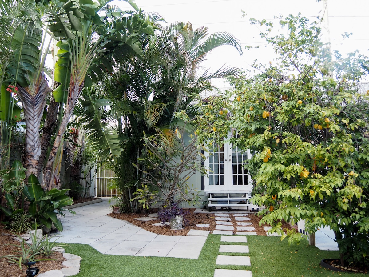 A lush garden walkway is visible, framed by vibrant tropical plants and trees. A stepping stone path leads to a clear backdrop of a light-colored house with multiple French doors. A bench rests among the greenery, inviting relaxation in the outdoor space.