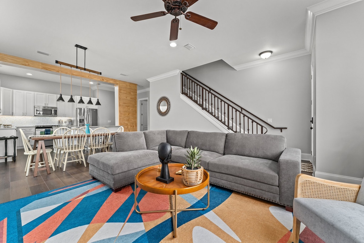 A spacious living area is highlighted by a large sectional sofa and a circular coffee table featuring a small plant. The room is adorned with a colorful area rug, and a dining table is visible in the background. A staircase leads to the second floor.