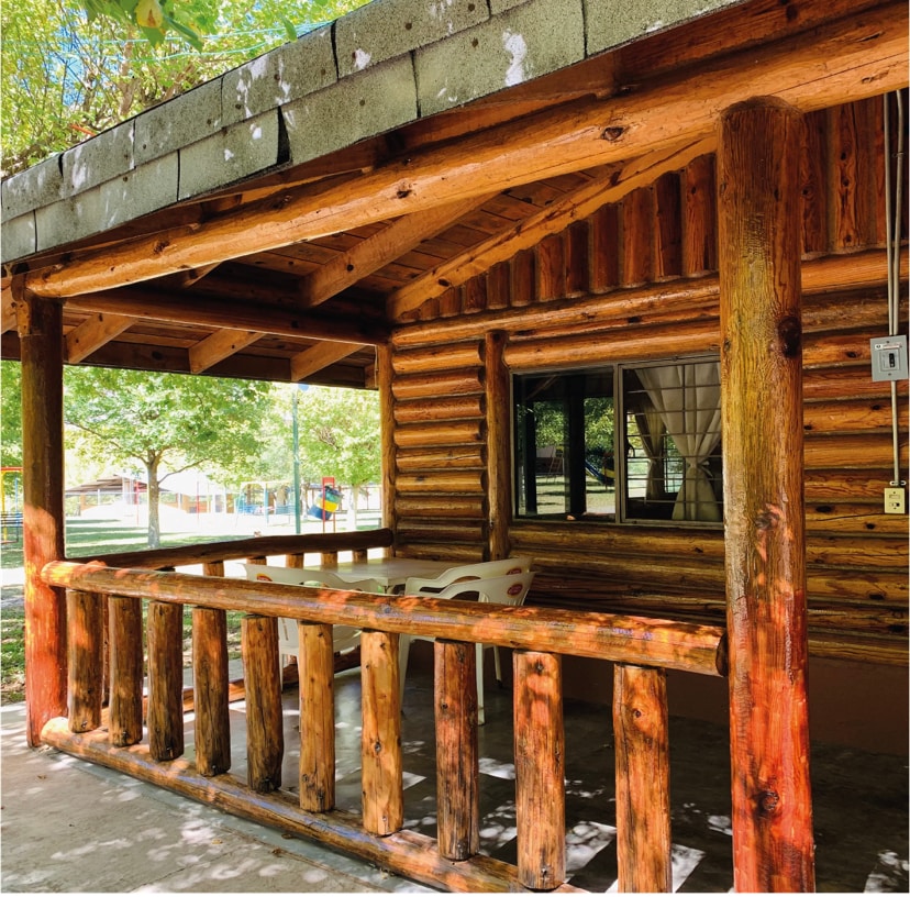 A rustic exterior view of a cabin is shown, featuring natural wood logs and a spacious porch. The area includes a railing made of thick wooden posts, with light filtering through the overhead structure. Large windows allow visibility to the surroundings, enhancing the connection to nature.