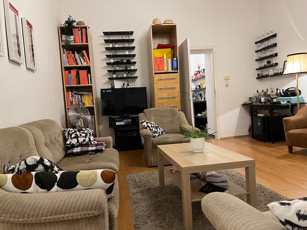 A comfortable living area features two light brown sofas, interspersed with colorful cushions. A coffee table sits centrally on a soft rug. In the background, a wooden bookshelf displays books, while a TV and a small bar area are visible, enhancing the inviting space.