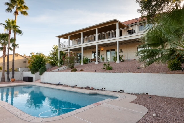 An outdoor view captures a large swimming pool framed by a landscaped yard and palm trees. The two-story home features a broad balcony and expansive deck space above, providing outdoor seating areas and access to various plants. A serene ambiance is highlighted by the clear sky.