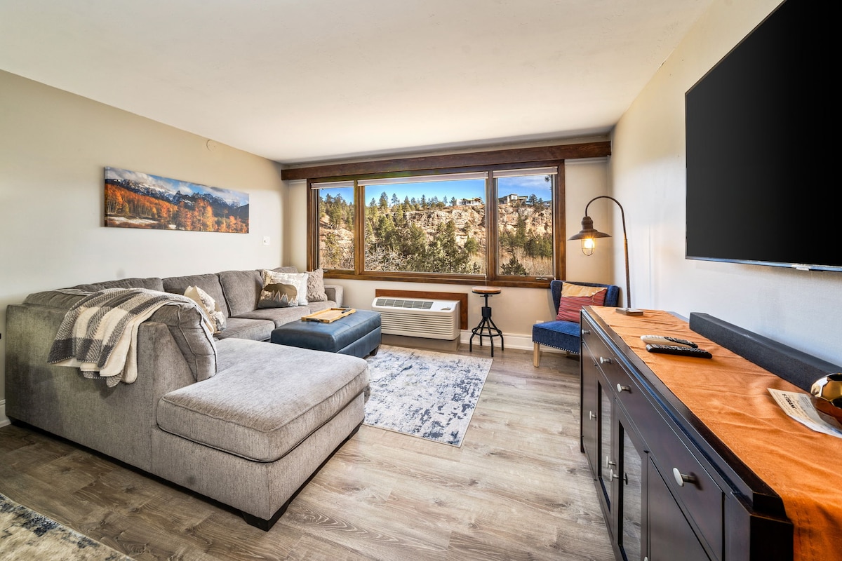 A spacious living area features a large sectional sofa, inviting a relaxed atmosphere. Large windows provide natural light and showcase views of the surrounding landscape. A TV is mounted on the wall, and a wooden console table adds functionality to the space.