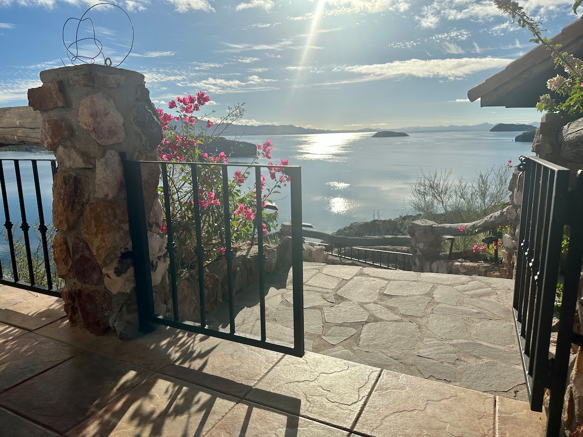 A sunlit stone patio offers a glimpse of the ocean views. Colorful flowering plants frame the entrance, with wrought iron railings leading towards the tranquil water. The smooth stone pathway extends into the landscape, where distant islands can be seen across the bay.
