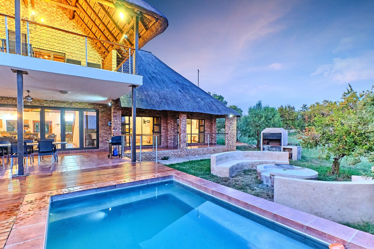 A spacious outdoor area features a private swimming pool surrounded by a wooden deck. Nearby, a built-in braai area is complemented by seating arranged around a stone firepit, while the lodge's brick exterior and thatched roof create a welcoming ambiance in the natural setting.