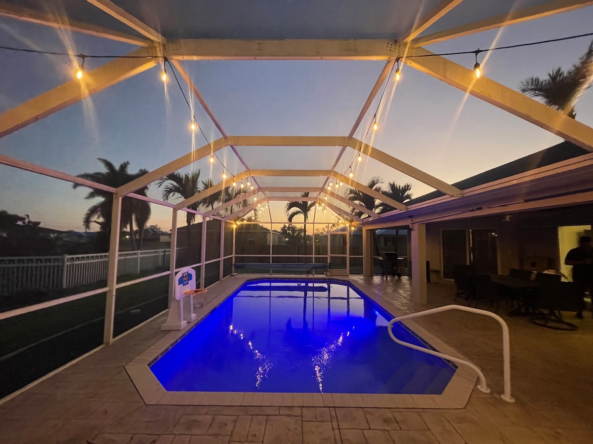 An outdoor space features a beautifully illuminated pool, reflecting shades of blue. The screened-in lanai is accented by string lights overhead, creating a serene evening ambiance. Palm trees are visible in the background, enhancing the tropical setting.