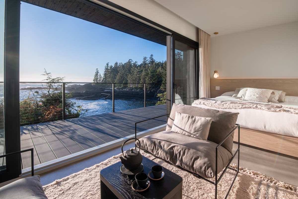 A spacious room features a large sliding glass door that provides an unobstructed view of the ocean and coastline. A comfortable bed is positioned nearby, with a cozy seating area that includes a low table and tea set, enhancing relaxation with natural light flooding the space.