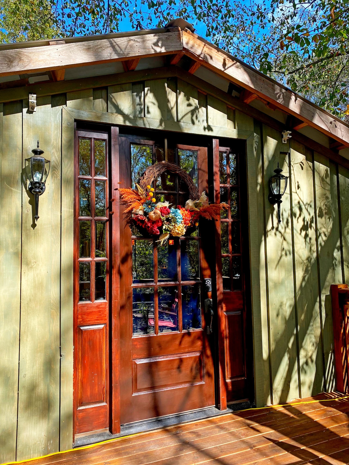 The entrance features a wooden door adorned with a floral wreath, complemented by large glass panels. Flanking the door, two wall-mounted lights provide illumination. Sunlight filters through surrounding trees, casting intricate shadows on the wooden deck.