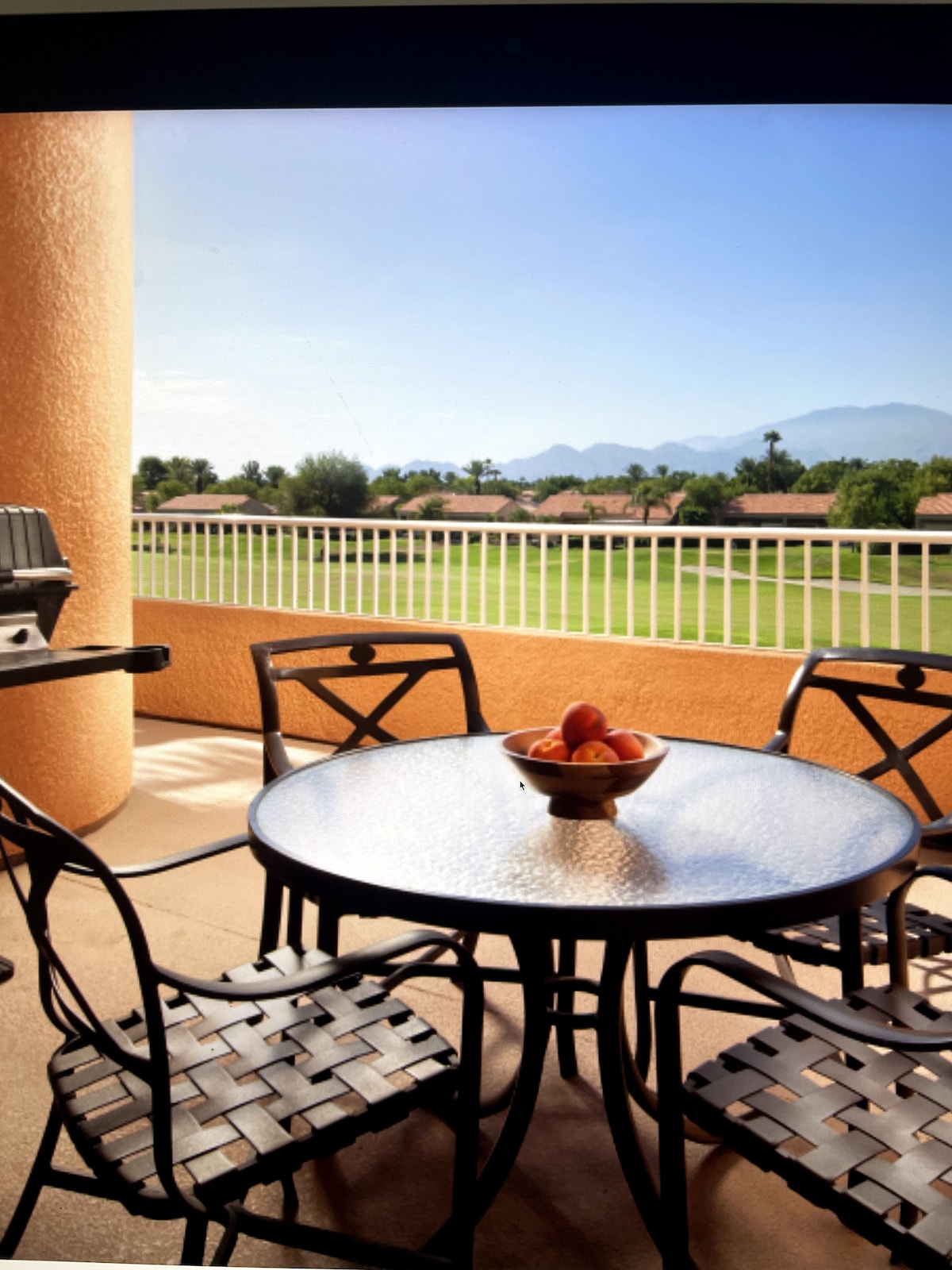 A private balcony features a round table surrounded by four metal chairs, with a bowl of fruit at the center. The backdrop includes a view of lush greenery and distant mountains, complemented by soft sunlight illuminating the area.