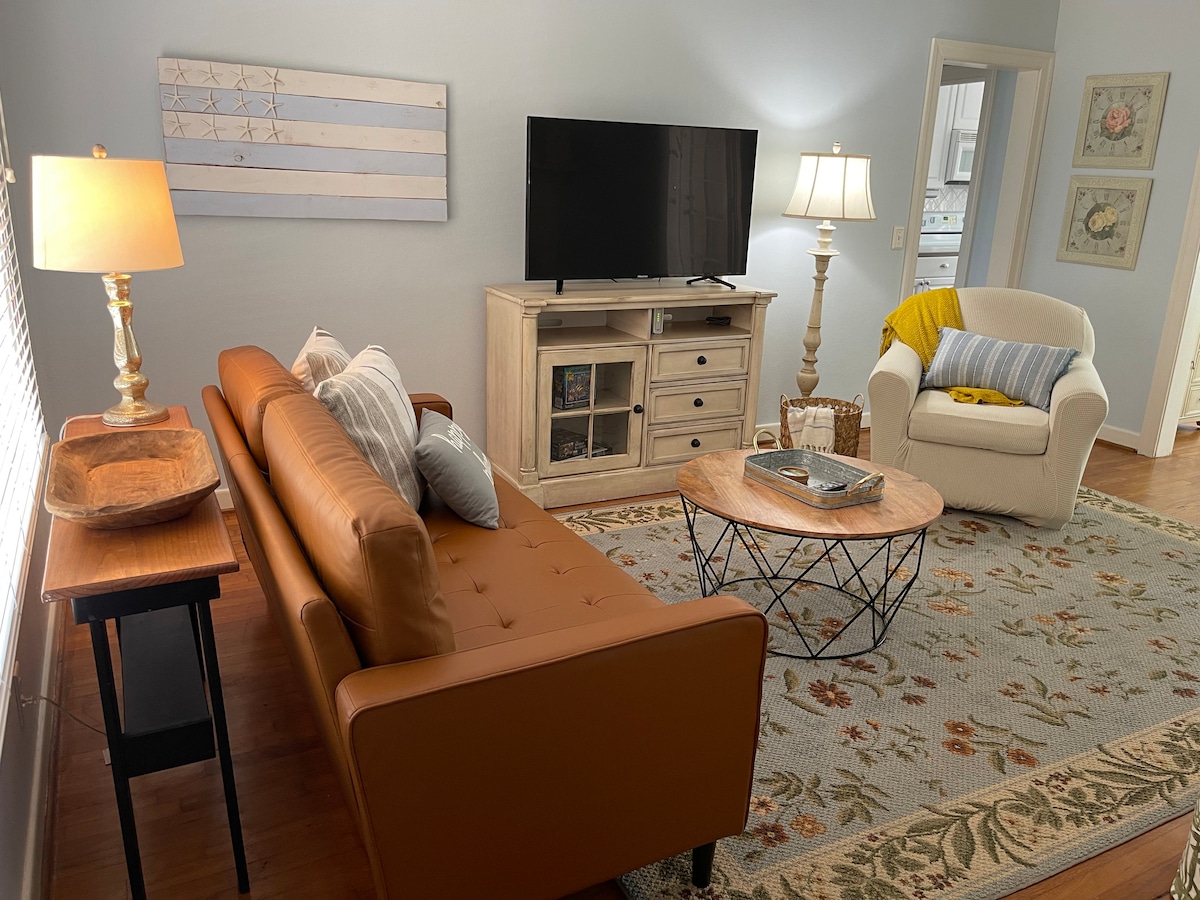 A comfortable living area features a brown leather sofa and a cozy armchair. Natural light brightens the space, highlighting a wooden coffee table with decorative items. A television is placed on a light-colored, multi-drawer console across from the seating, and an area rug adds warmth to the flooring.