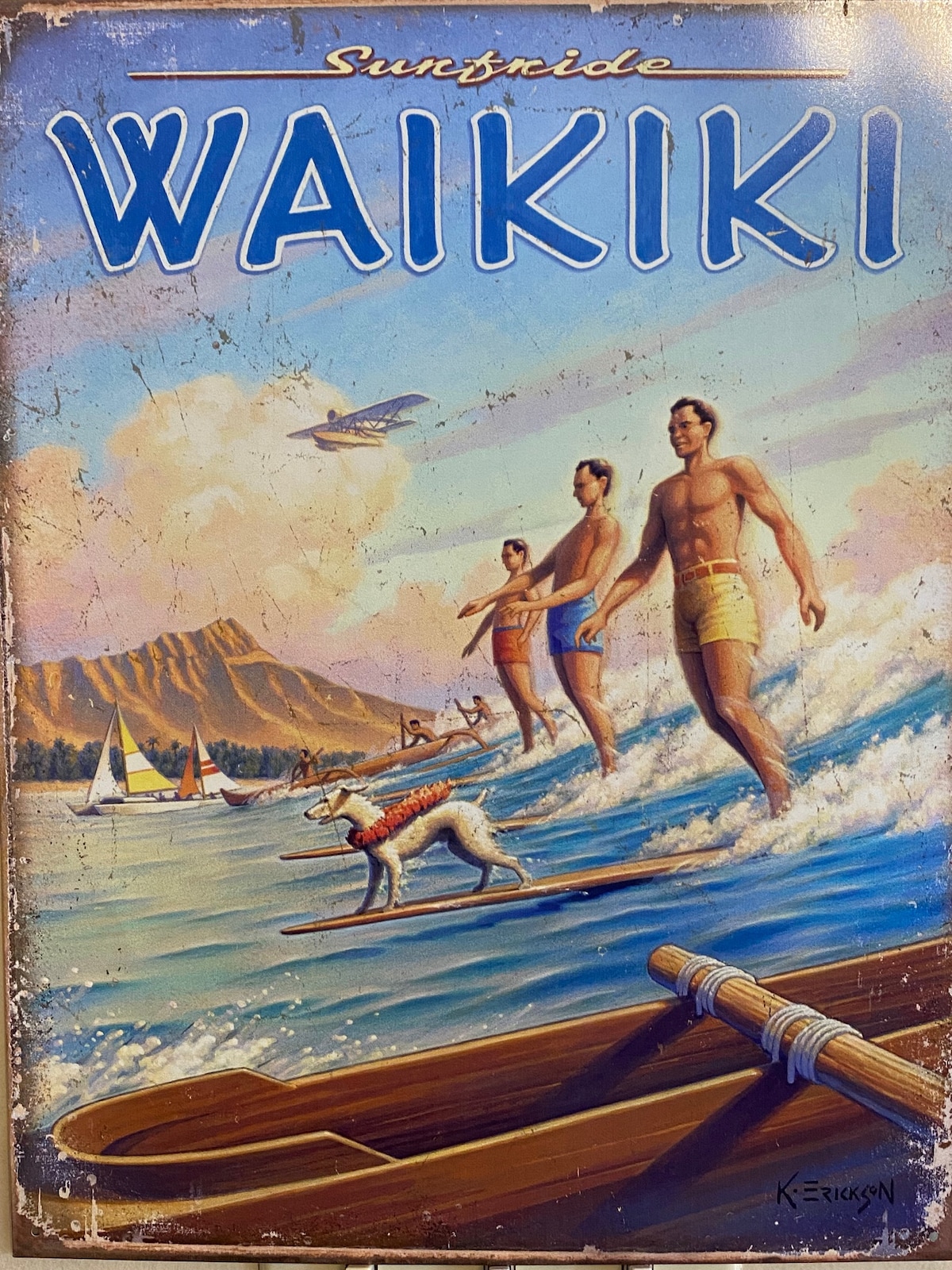 An artistic retro depiction of surfers riding waves at Waikiki, featuring three male figures in colorful swim trunks and a dog on a surfboard. In the background, sailboats and a scenic mountain landscape are visible under a clear sky.